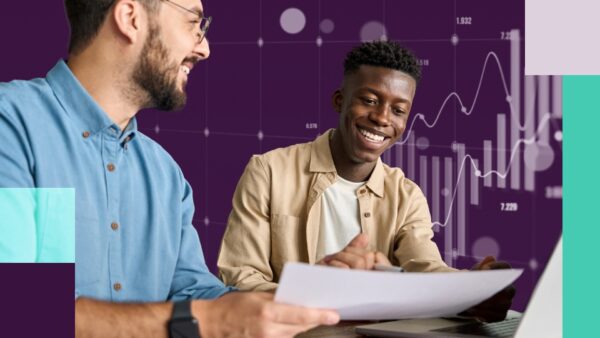 Two men smiling while discussing something on a piece of paper. There are finance graphs in the background.
